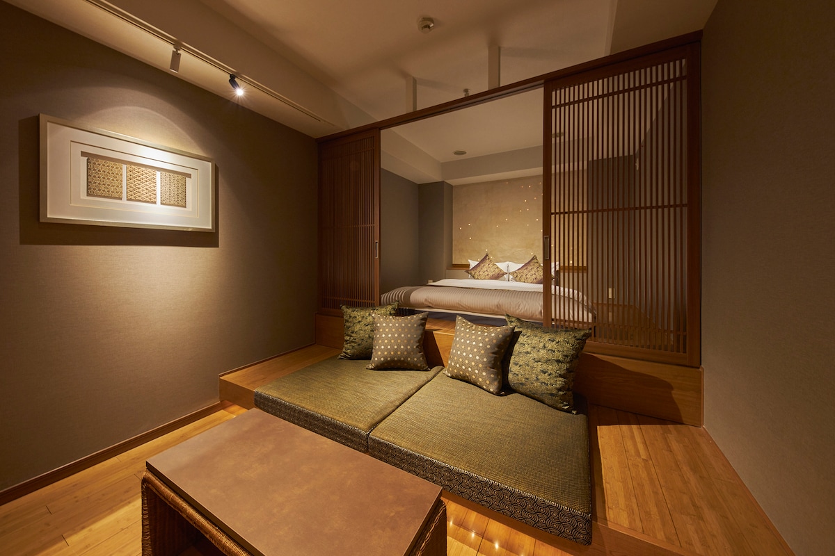A spacious room features a cozy sitting area with cushioned seats and a low wooden table. Behind a partition, a comfortable bed is seen, complemented by soft lighting and subtle wall decor, enhancing the serene atmosphere.