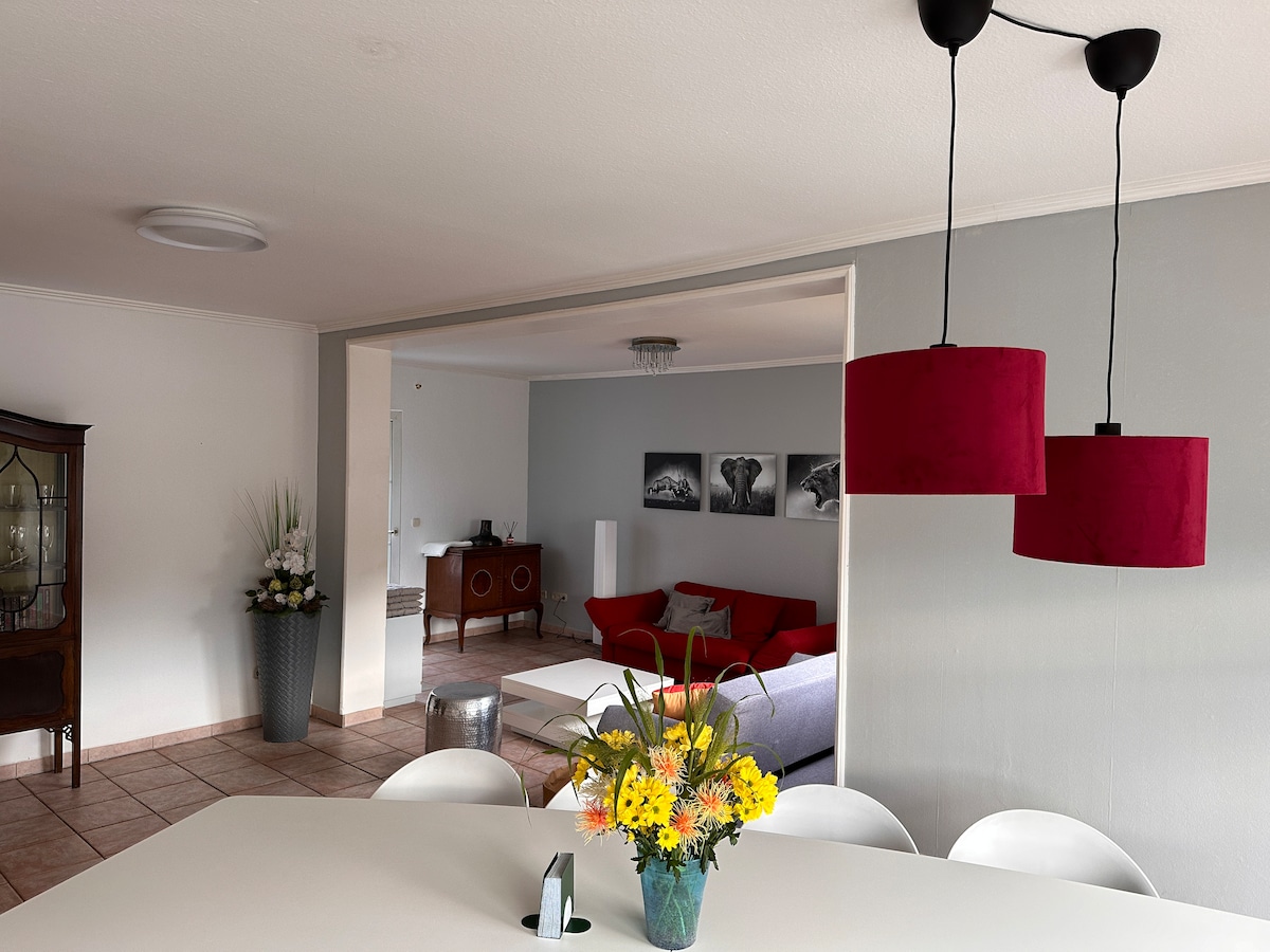 An open dining area is seen, featuring a round table and white chairs. A simple vase with vibrant yellow flowers is centerpiece. Two red pendant lights hang above the table, while a spacious living room is visible in the background, with a comfortable red sofa and wall art.