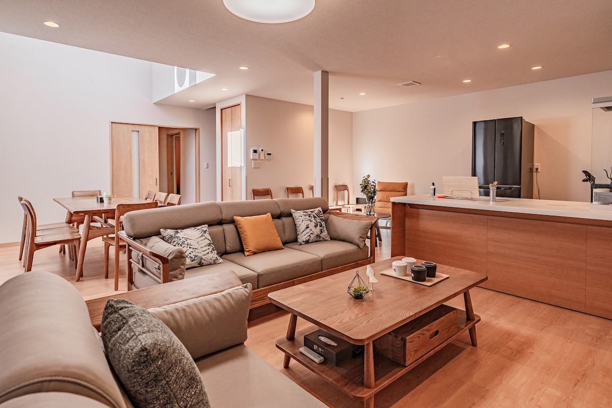 An open-concept living area features a light-filled space with soft seating on one side and a dining area on the other. A wooden coffee table holds decorative items, while modern appliances are visible in the kitchen area, contributing to a welcoming environment.
