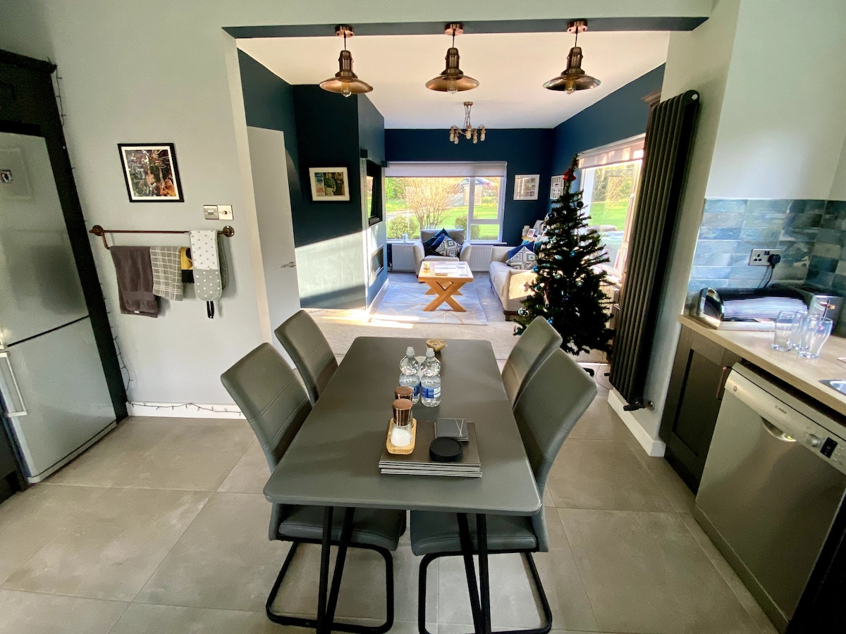 A modern dining area features a rectangular table surrounded by six grey chairs. The space connects to a bright living room visible through an open doorway, where natural light highlights a cozy seating arrangement and a Christmas tree stands in the corner.