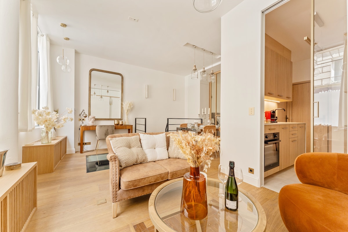 A bright living area showcases a comfortable sofa adorned with decorative cushions, accompanied by a stylish armchair. A wooden dining table is visible, along with a modern kitchen in the background. Natural light floods the space through large windows, creating an open and inviting ambiance.