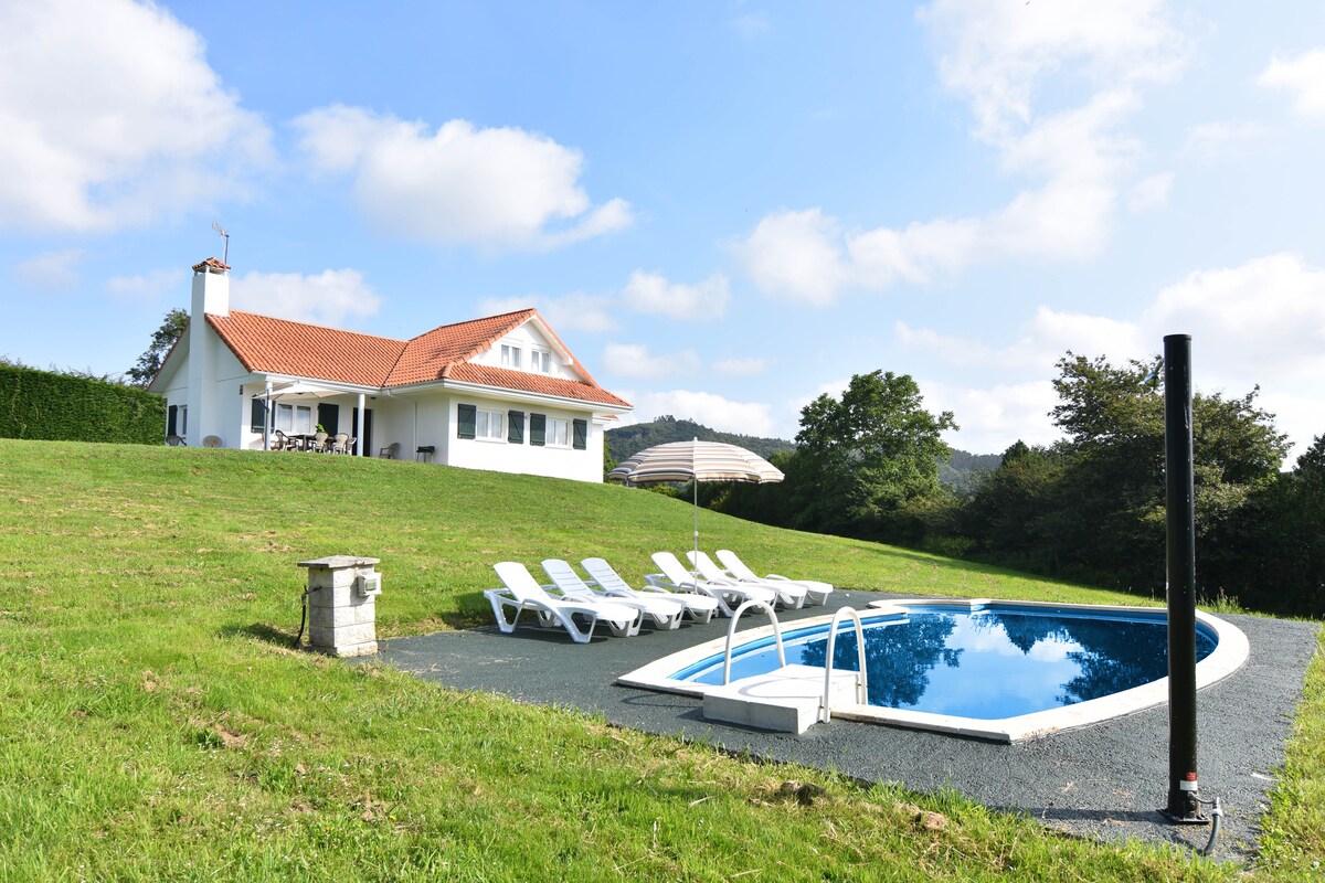A spacious detached holiday home is featured against a backdrop of lush greenery. The private swimming pool, surrounded by comfortable sun loungers, is complemented by a shaded area provided by an umbrella. The home presents a welcoming façade with a classic roof and expansive lawn.