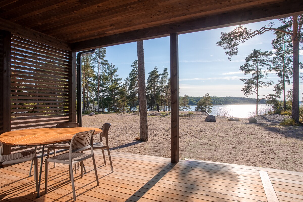 A covered terrace extends from the cabin, featuring a round wooden dining table surrounded by metal chairs. The view showcases a sandy beach and a shimmering lake, framed by tall trees under a clear blue sky.