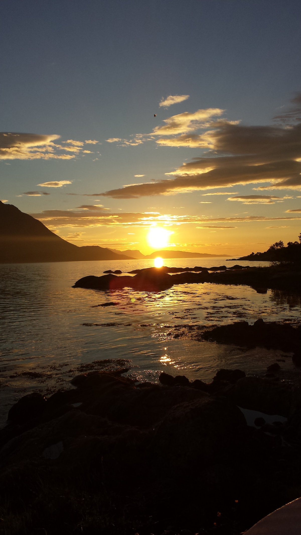 A serene sunset is captured over the fjord, with warm hues reflecting off the shimmering water. Silhouettes of distant mountains frame the scene, while rocky elements in the foreground add texture to the tranquil landscape.