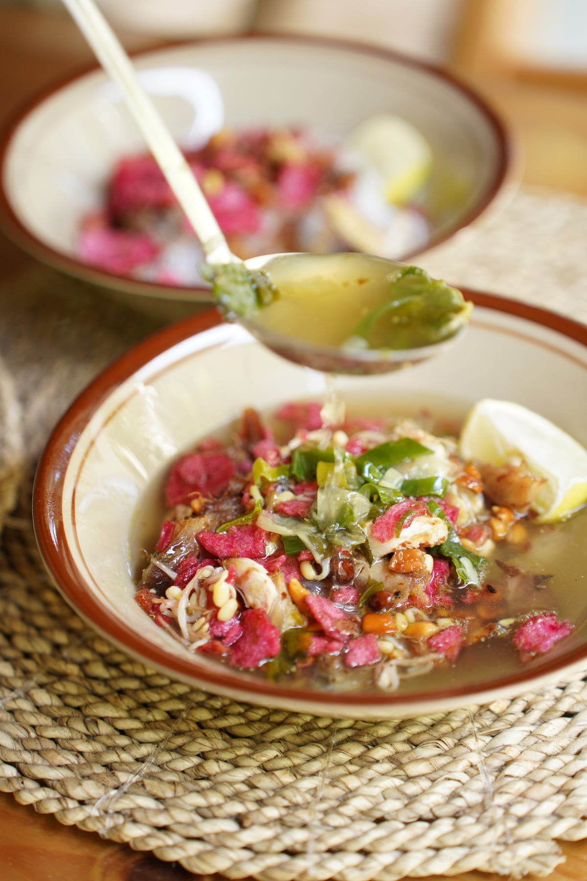 Two bowls of soup are presented on a woven mat. Each bowl features a clear broth filled with various vegetables and colorful toppings. A spoon, partially submerged, reveals herb garnishes and small pieces of meat, complemented by a wedge of lime on the side.