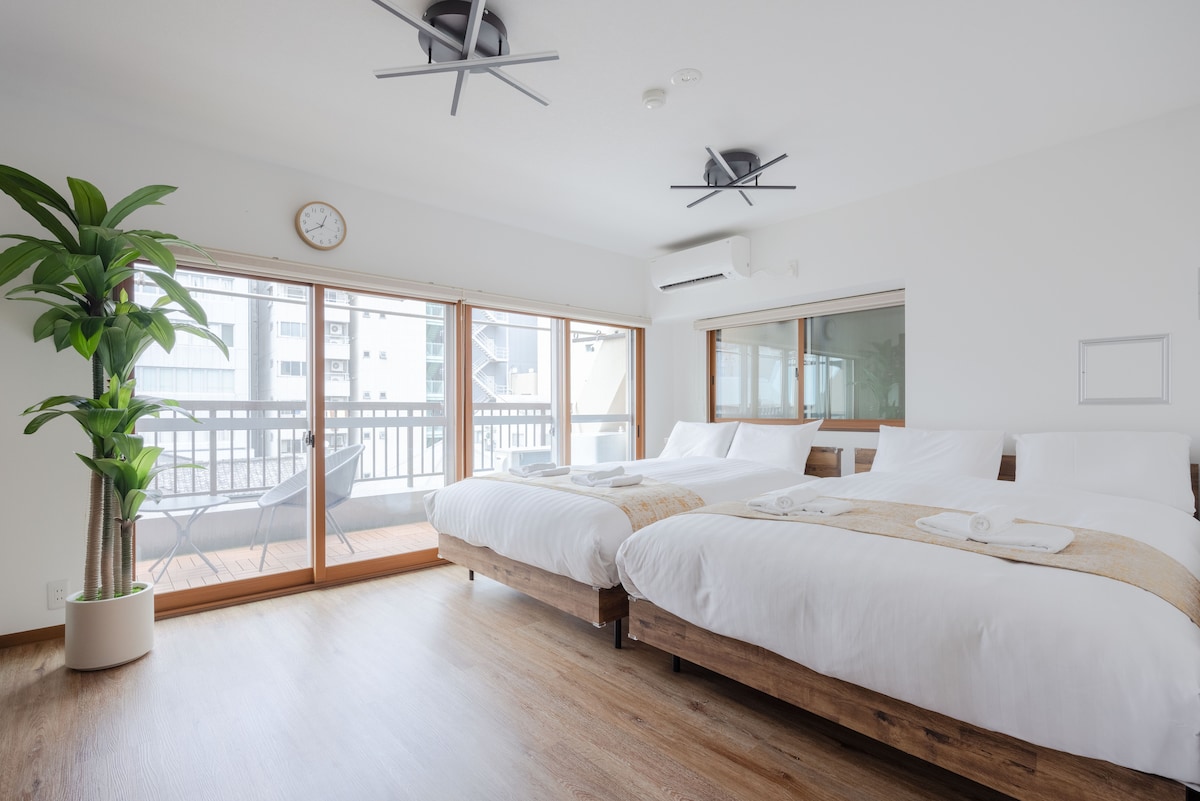 A bright sleeping area is furnished with two spacious double beds, featuring crisp white linens. Large windows allow natural light to fill the room, while a small balcony is accessible. A potted plant adds a touch of greenery to the neutral decor.
