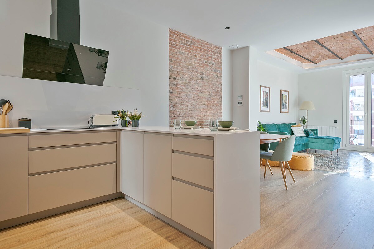 A modern kitchen area features light-colored cabinetry with a central island, complemented by a sleek black backsplash. In the background, a living space is visible, showcasing a green sofa and dining area, providing a sense of openness and cohesive design.