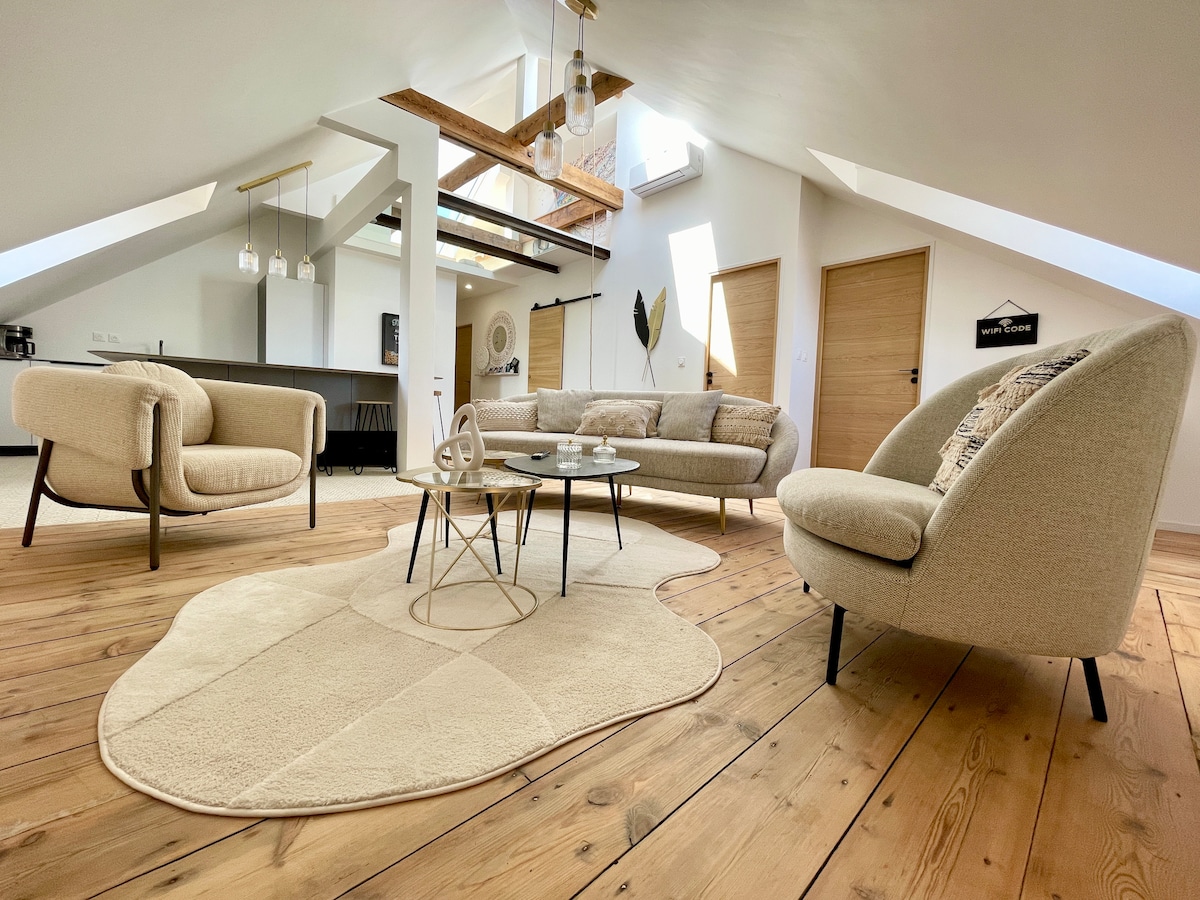 An airy living space is presented with natural wood flooring and sloped ceilings. A neutral-toned rug sits beneath a modern coffee table, surrounded by two distinctive chairs and a plush sofa. Warm light filters through windows, enhancing the inviting atmosphere.