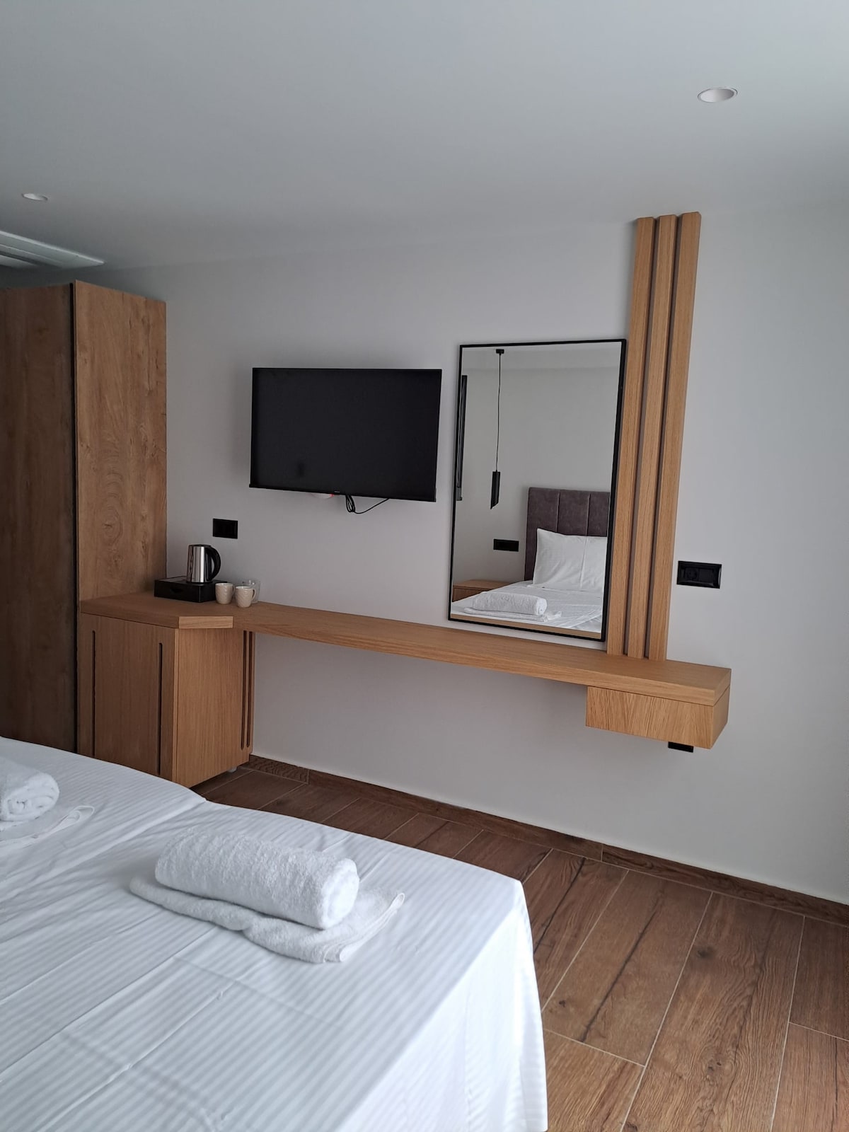 A modern guest room is depicted, featuring a spacious bed with crisp linens. A wooden console with a mounted mirror and flat-screen television is visible, along with a kettle and two cups. Light hardwood flooring complements the clean design.