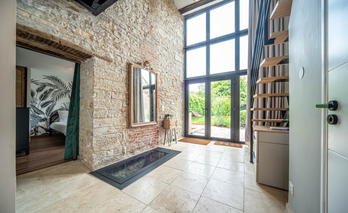 A bright entryway features a large window wall allowing natural light to fill the space. Stone walls complement a mirrored furniture piece, and a staircase leads to an upper level. A serene view of the outdoors is visible through the glass doors.