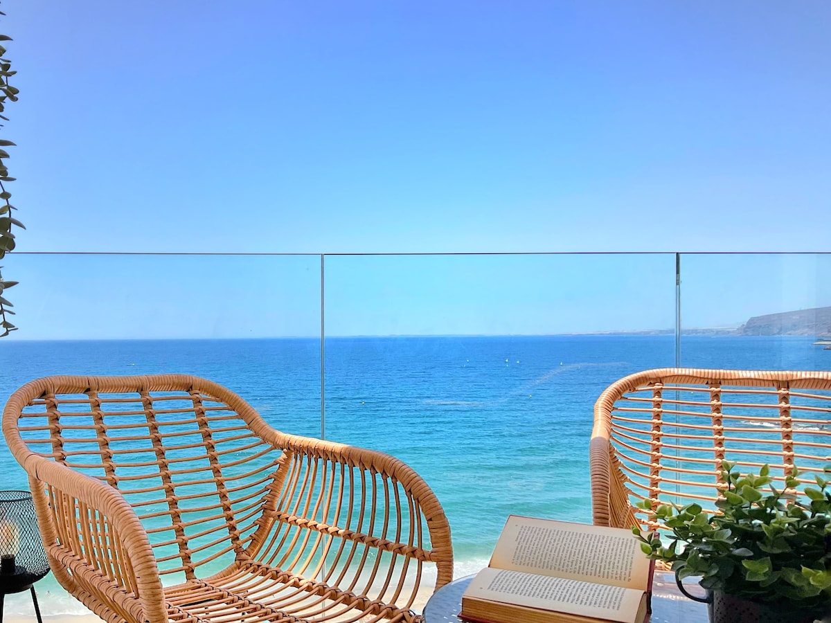 A terrace features two woven chairs positioned beside a small table, overlooking a clear blue sea. A book lies open on the table, and a potted plant adds a touch of greenery. The sky above is bright and clear, enhancing the tranquil setting.