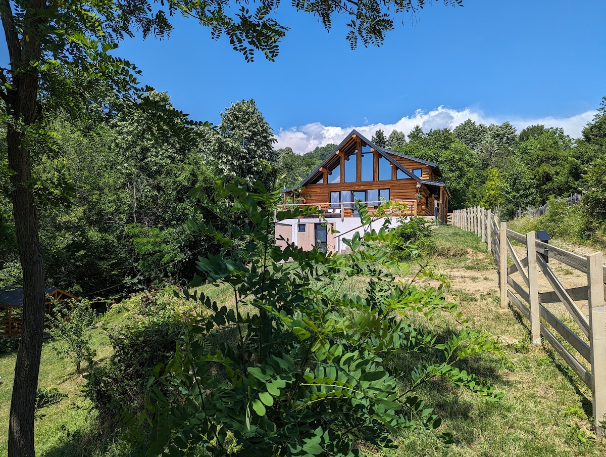 A wooden chalet emerges amidst lush greenery, featuring large windows that invite natural light. A wooden fence lines the property, with trees framing the view. The clear blue sky complements the serene landscape surrounding the home.