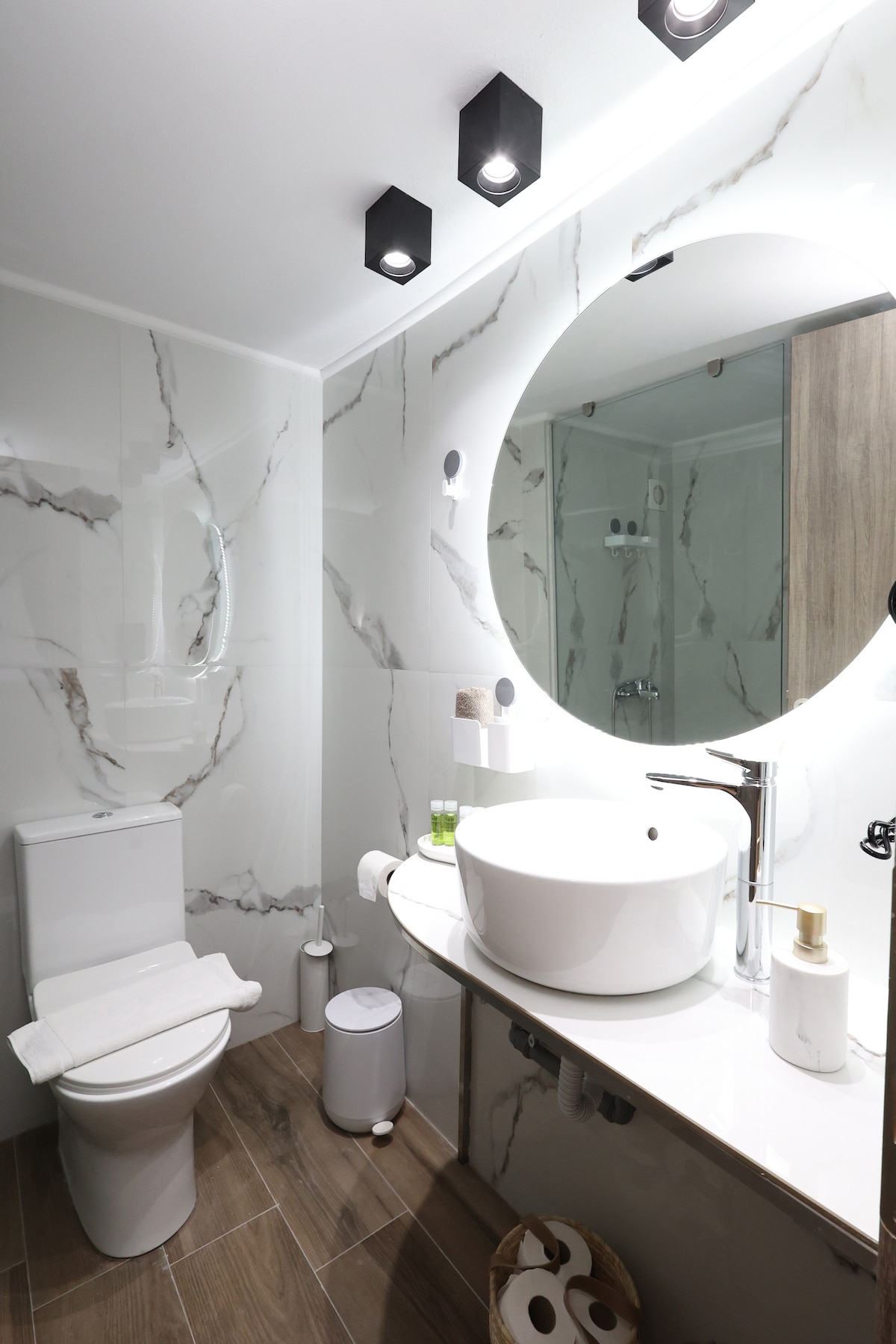 A modern bathroom features elegant marble walls and sleek fixtures. A round mirror illuminates the space above a stylish vessel sink. A toilet and a small waste bin are neatly positioned, while beige towels are arranged on the countertop, enhancing the room's functional design.