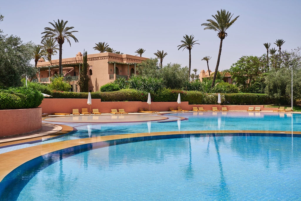 A serene swimming pool stretches across the image, reflecting the clear sky above. Sun loungers are arranged around the pool, partially shaded by white umbrellas. Lush greenery and palm trees create a relaxed atmosphere, while the surrounding architecture hints at traditional Moroccan design.