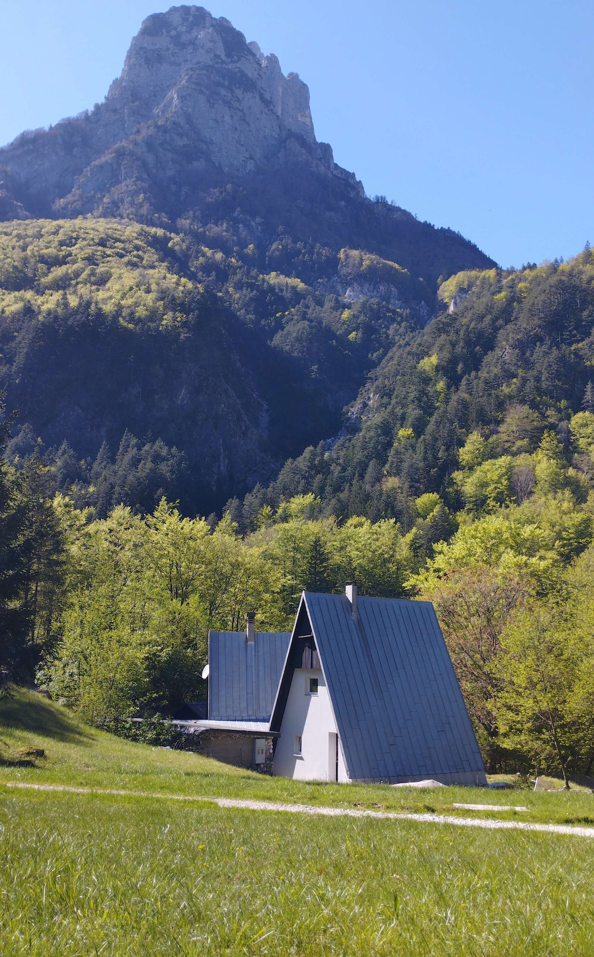 A mountain cabin is situated in a lush green landscape, surrounded by tall trees and steep mountain slopes. The A-frame structure features a sloped metal roof, blending with the natural scenery. The clear blue sky creates a bright backdrop, enhancing the serene environment.