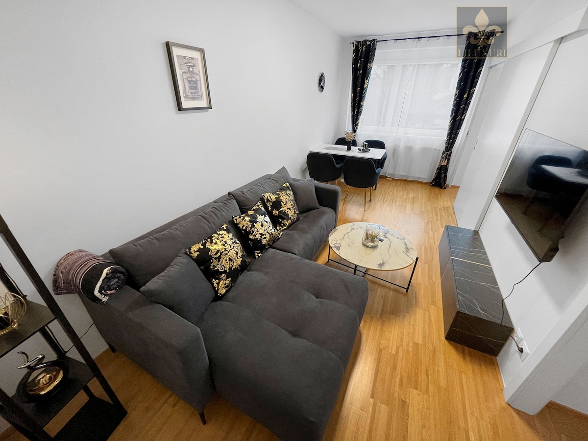 A spacious living area is presented with a dark gray sectional sofa adorned with decorative pillows. A round coffee table is placed in front, and a dining area is seen with two chairs and a table. Natural light filters through the large window, complemented by dark curtains.