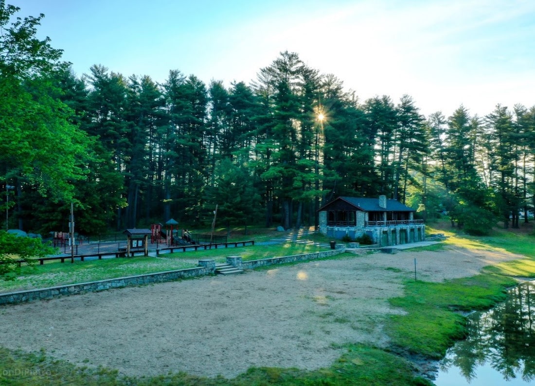 A serene landscape showcases a playground adjacent to a rustic structure nestled among tall trees. Sunlight filters through the branches, casting gentle rays over the fenced area. The grassy ground appears dry and sandy, indicating a transition in the environment.