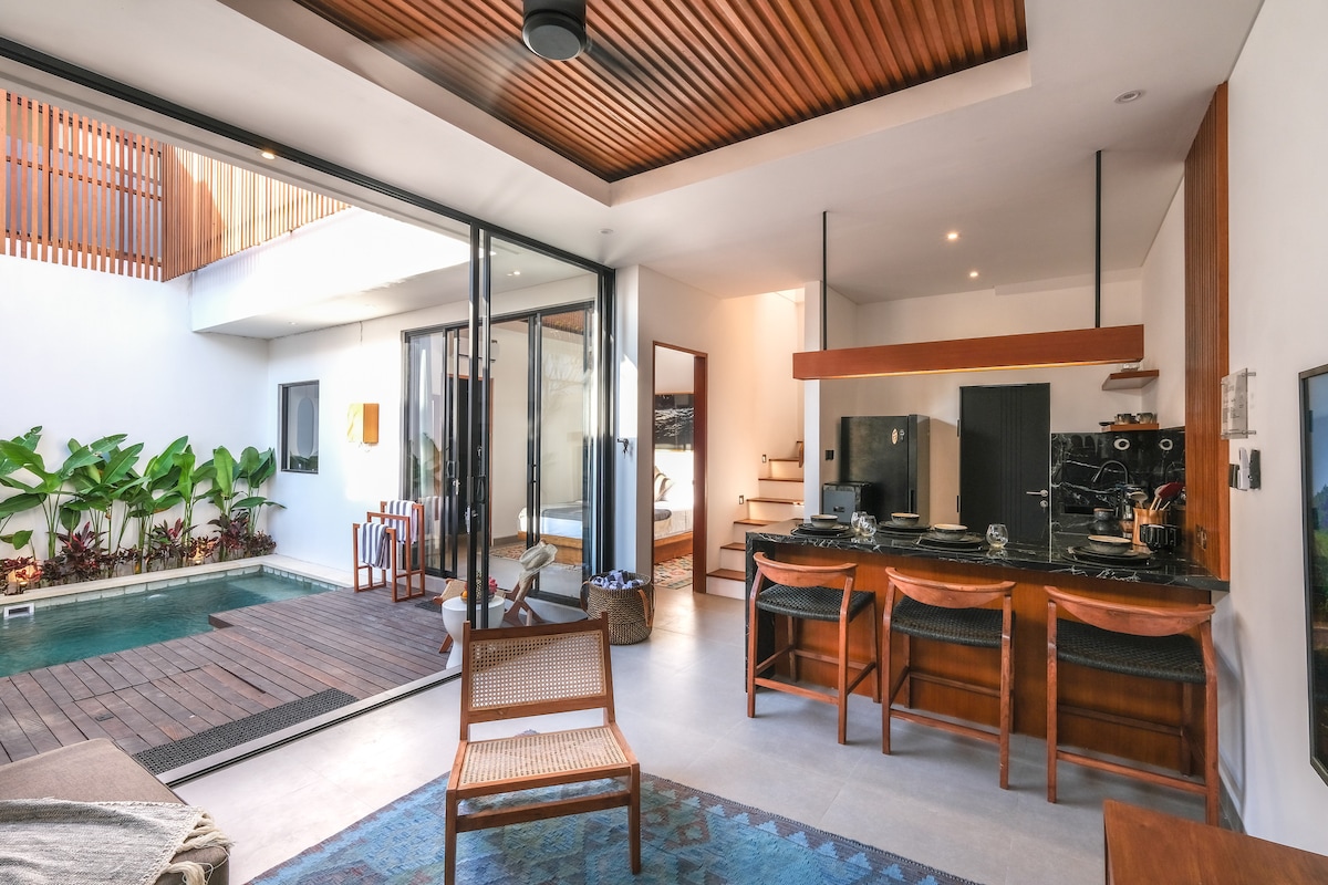 An open-plan living area showcases a comfortable seating arrangement and a kitchen with modern appliances. Large glass doors provide views of the private pool and surrounding greenery, enhancing the indoor-outdoor flow, while natural light brightens the space.
