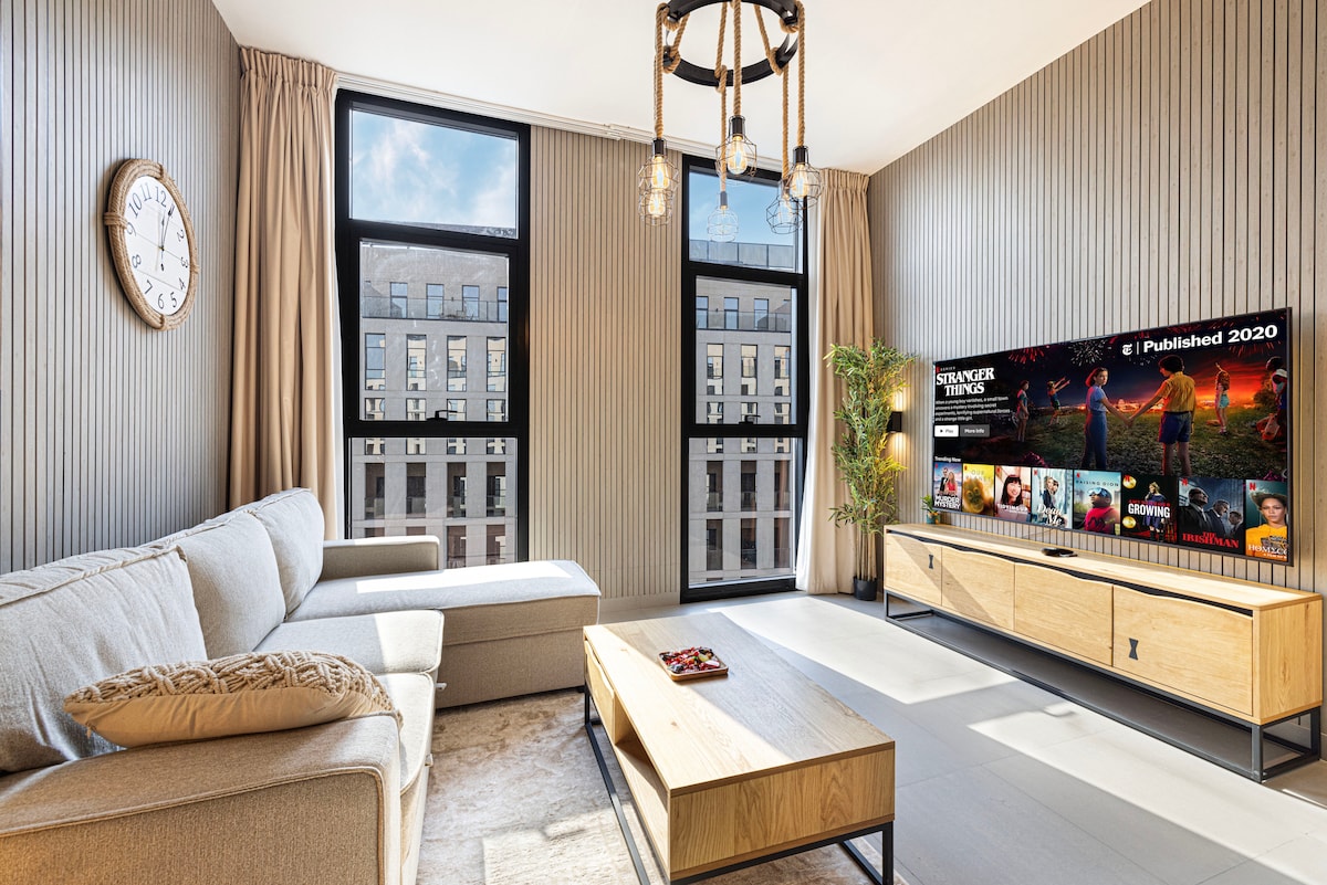 A modern lounge area features a comfortable sofa with a chaise and a wooden coffee table. Large windows allow natural light to fill the space, showcasing a stylish entertainment unit with a flat-screen TV displaying Netflix selections.