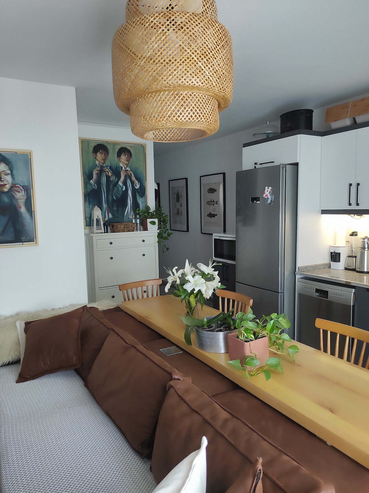 A modern kitchen and dining area are featured, with a wooden table centered among green plants. A woven pendant light hangs from the ceiling above. A refrigerator and oven are visible in the background, complemented by artwork on the walls. Neutral tones create a calm atmosphere.