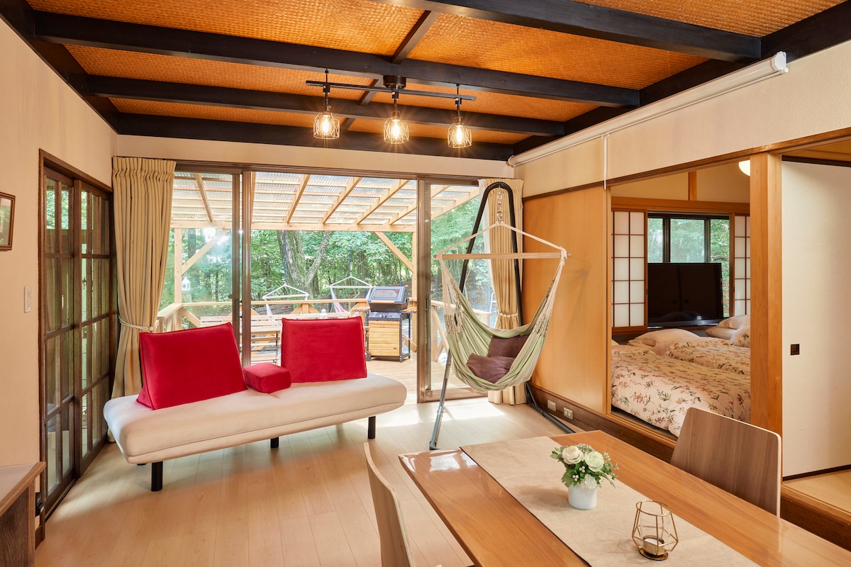 A spacious living area features a white sofa adorned with red cushions, complemented by a dining table. Large glass doors open to a wooden deck, allowing natural light to illuminate the space. A cozy hammock is suspended nearby, adding a relaxed ambiance.