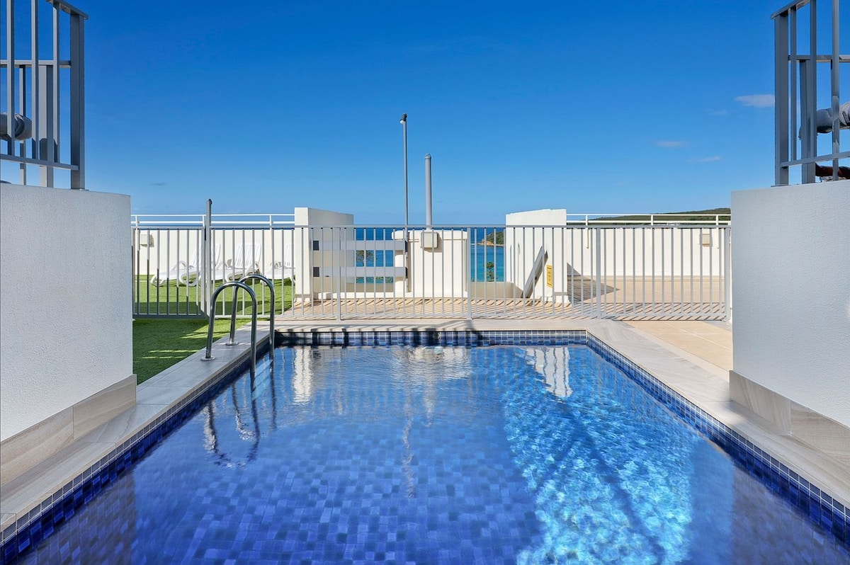 A rooftop pool offers a clear view of the sky and surrounding area. Sunlight reflects off the water's surface, highlighting the blue tiles of the pool. Lounge chairs are positioned nearby for relaxation, creating an inviting outdoor space.
