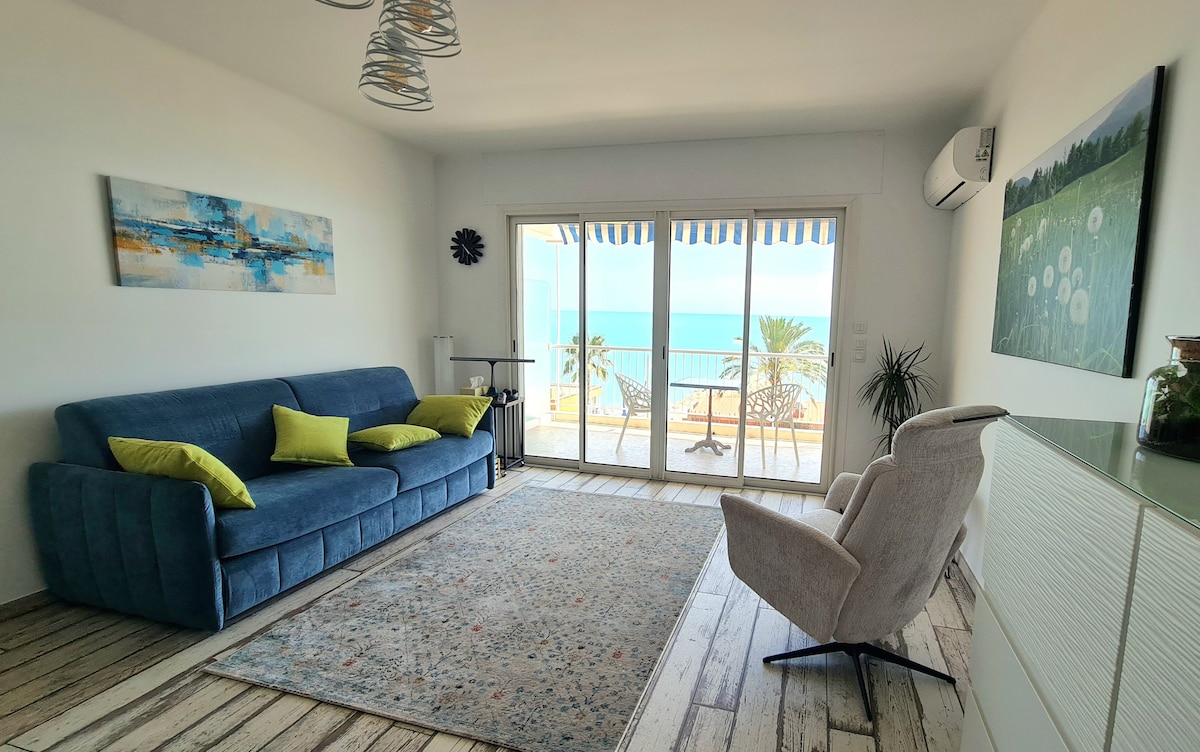 A spacious living area is shown, featuring a plush blue couch adorned with green cushions. Large glass doors open to a balcony, providing a view of the sea. A cozy armchair and a patterned rug enhance the inviting atmosphere, along with light wooden flooring.