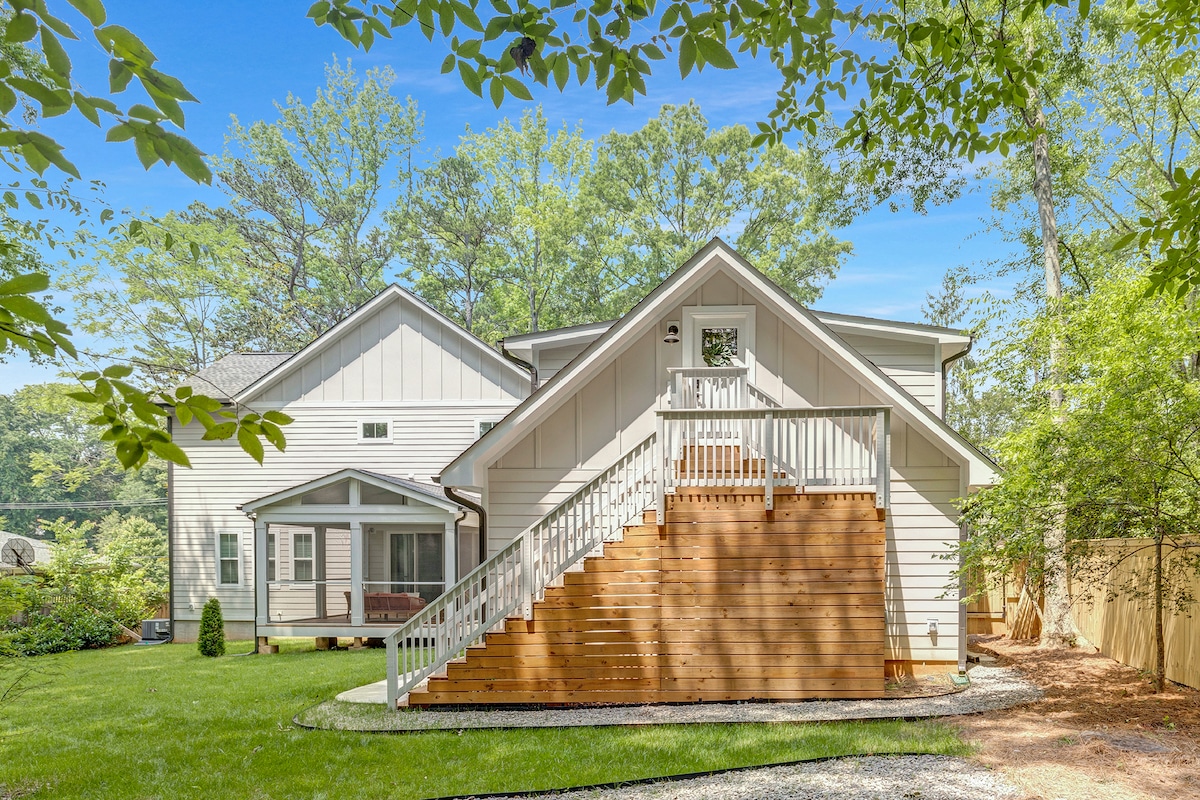 The image shows the exterior of a newly built guest house surrounded by lush greenery. Stairs lead up to a wooden deck, while a grassy area and trees create a serene environment. Natural light enhances the modern architectural design.