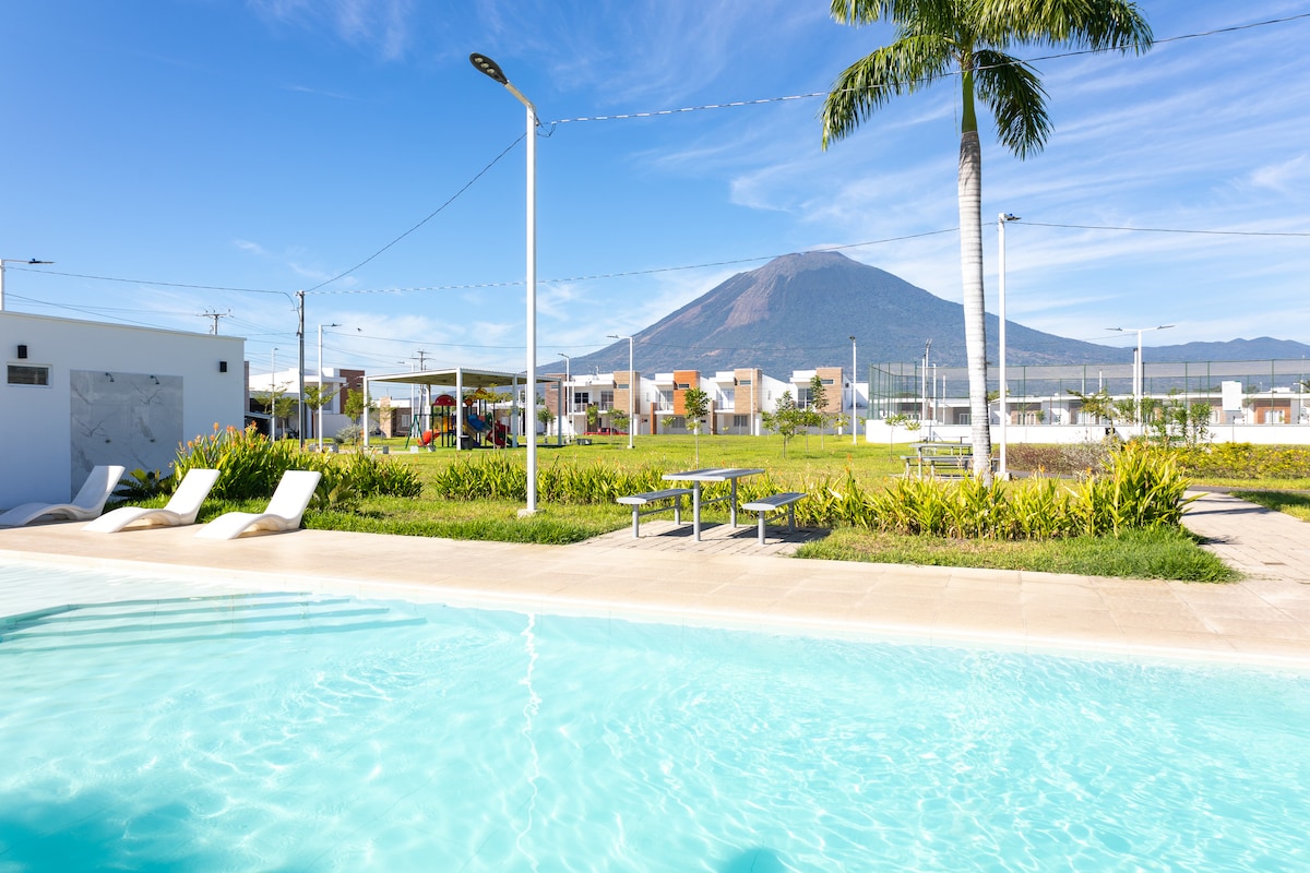 A tranquil pool area is framed by lush landscaping, with lounge chairs positioned by the water. In the background, a prominent mountain can be seen under a clear blue sky, contributing to a serene environment. Nearby, a grassy space provides room for outdoor activities.
