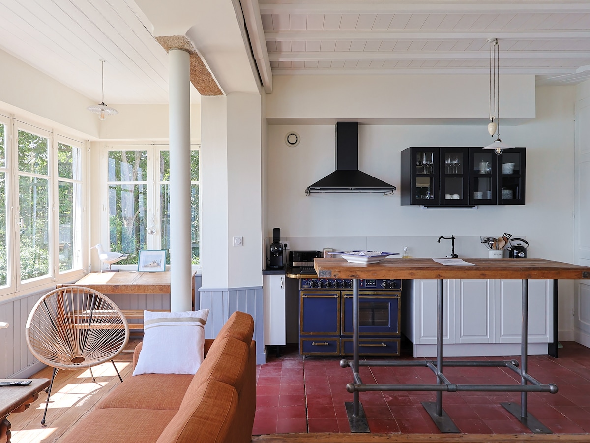 The spacious kitchen area features a wooden island with bar seating and modern appliances, including a blue oven and a black range hood. Large windows on three sides allow natural light to fill the space, with a cozy seating area visible nearby.