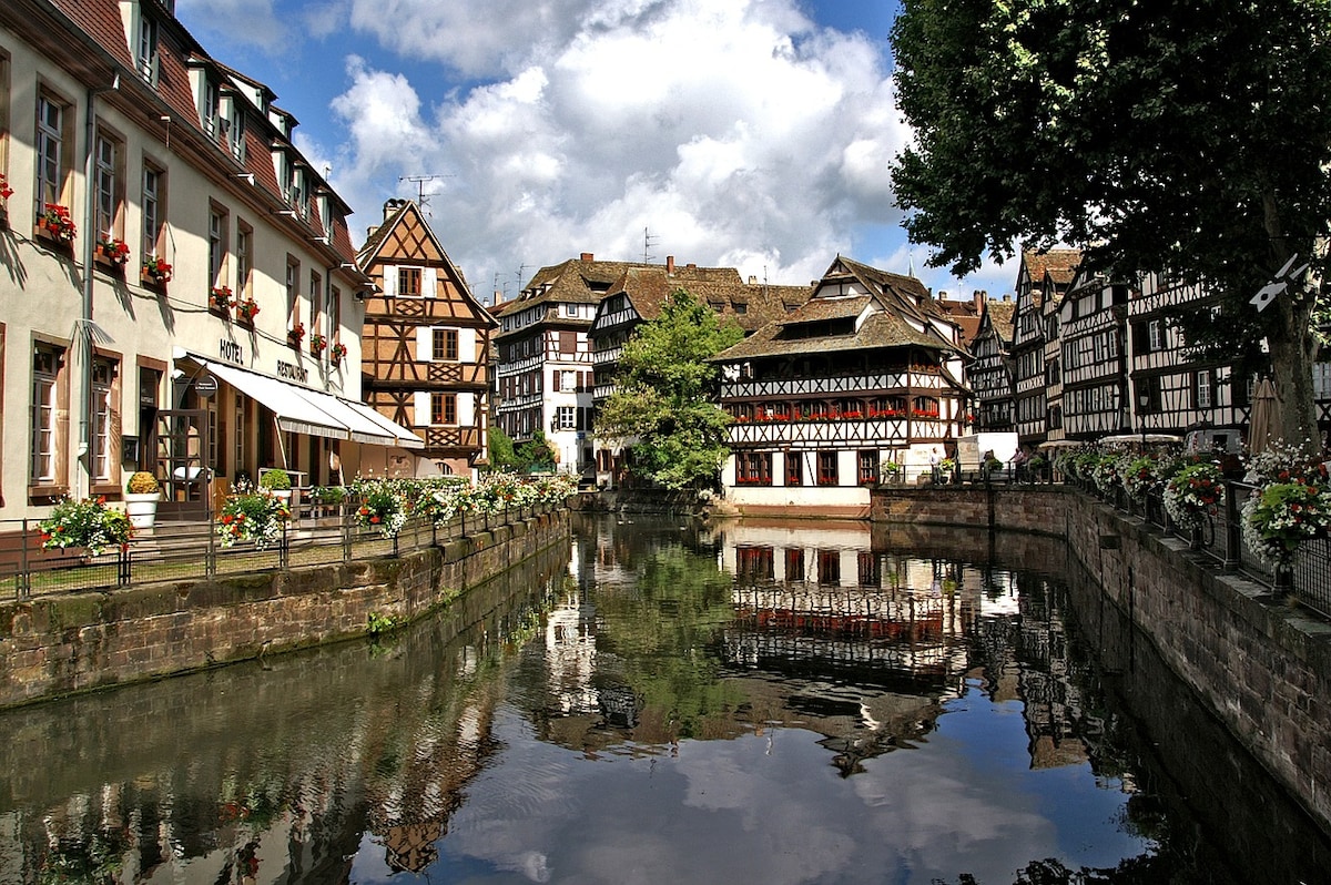 A scenic view of a picturesque canal lined with half-timbered houses, reflecting beautifully in the water. Flower boxes adorn the buildings, creating a charming ambiance. Lush green trees provide shade, while the sky is dotted with fluffy clouds, enhancing the serenity of this quaint area.