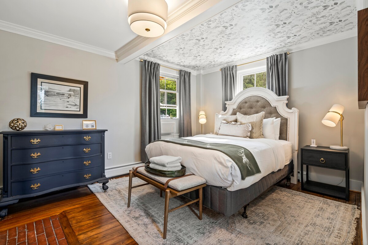 A well-appointed guest room is featured, showcasing a queen-sized bed with plush bedding. Natural light enters through two windows, and soft gray curtains frame each window. A dark wooden dresser and nightstand complement the space, while a woven rug adds warmth to the wooden floor.