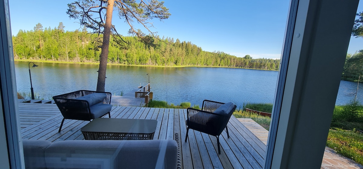 A view from inside the cabin showcases a spacious deck with two comfortable lounge chairs and a table facing a serene lake. The calm water reflects the surrounding greenery, with a tall tree positioned nearby. The bright sky enhances the tranquil atmosphere of the scene.