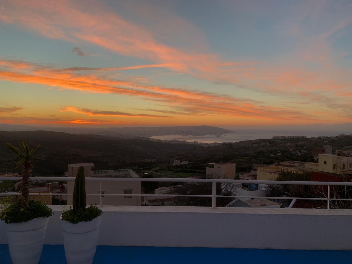 A spacious rooftop terrace is shown, featuring two large planters with greenery. In the background, a stunning sunset casts warm hues across the sky, while the bay of Tanger stretches out below, blending with the lush landscape. The scene offers a peaceful, expansive view.