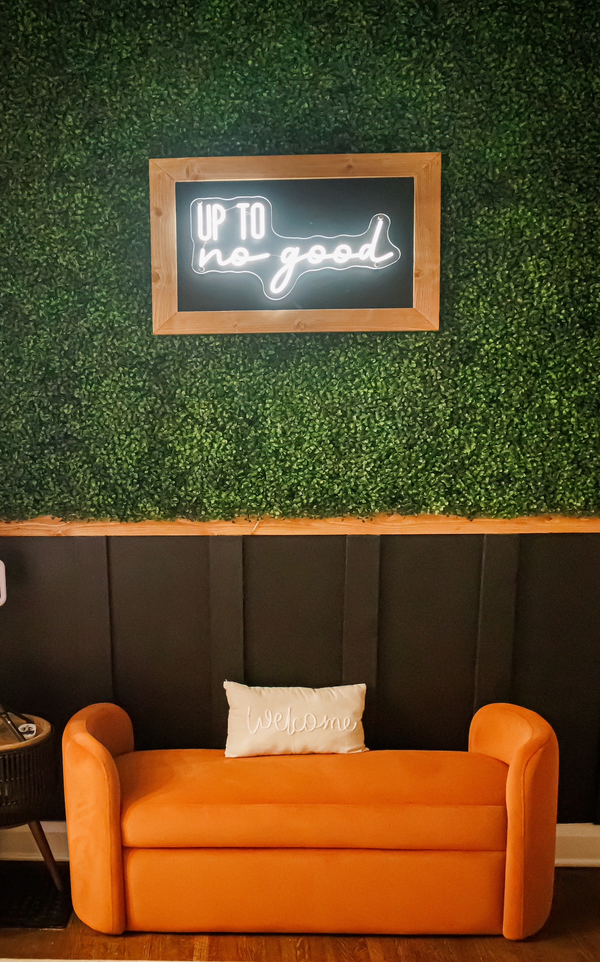 A cozy orange sofa is positioned against a green foliage wall, featuring a charming 'Welcome' pillow. Above the sofa, a wooden-framed neon sign reads 'Up to no good,' enhancing the playful atmosphere of the space.