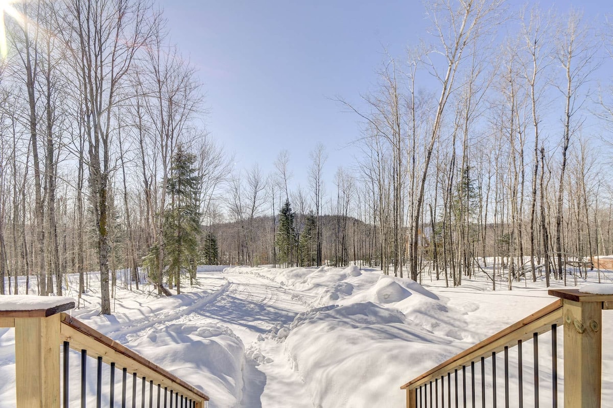 A snowy landscape is viewed from a wooden deck, with gentle slopes covered in fresh snow leading into a forested area. Bare trees stand tall under a clear blue sky, creating a serene winter atmosphere.