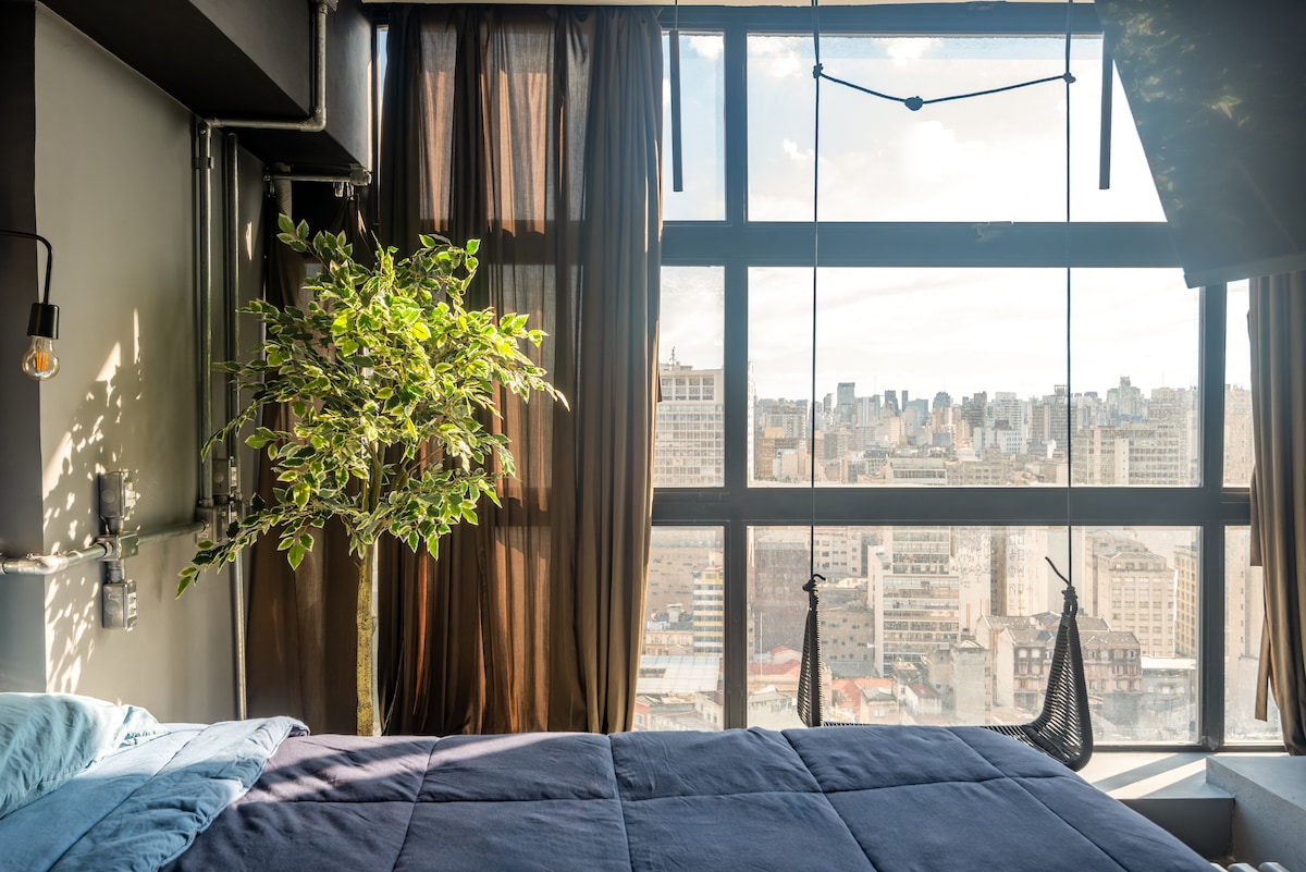 A spacious bedroom features a large window offering a panoramic view of the cityscape. A plant is placed near the window, complemented by warm curtains. A cozy bed with an inviting blanket is positioned near the window, creating a serene atmosphere.