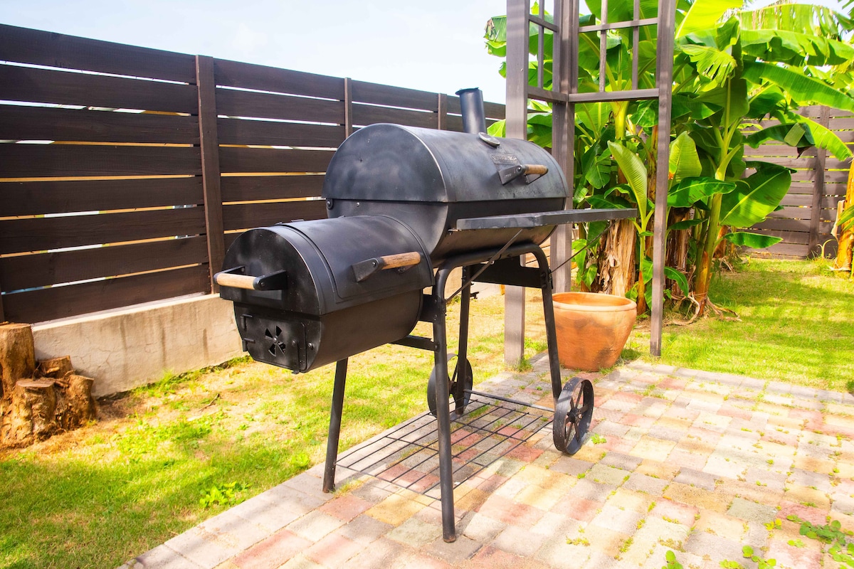 A black charcoal grill is positioned on a paved patio in a grassy area. The grill features a rounded body and a side shelf for food preparation. Surrounding plants and a wooden fence provide a natural and private setting.