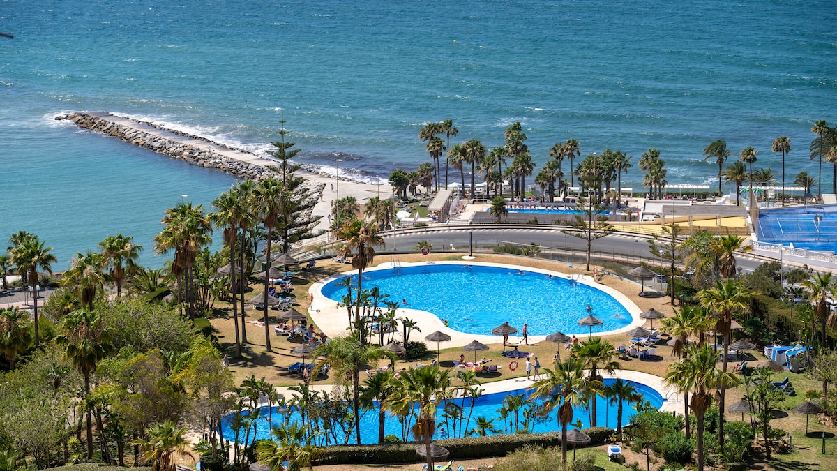 A spacious outdoor area includes two large swimming pools surrounded by palm trees. Sun loungers are arranged throughout the area, providing options for relaxation. The coastline is visible in the background, highlighting the proximity of the beach and the vibrant blue ocean.