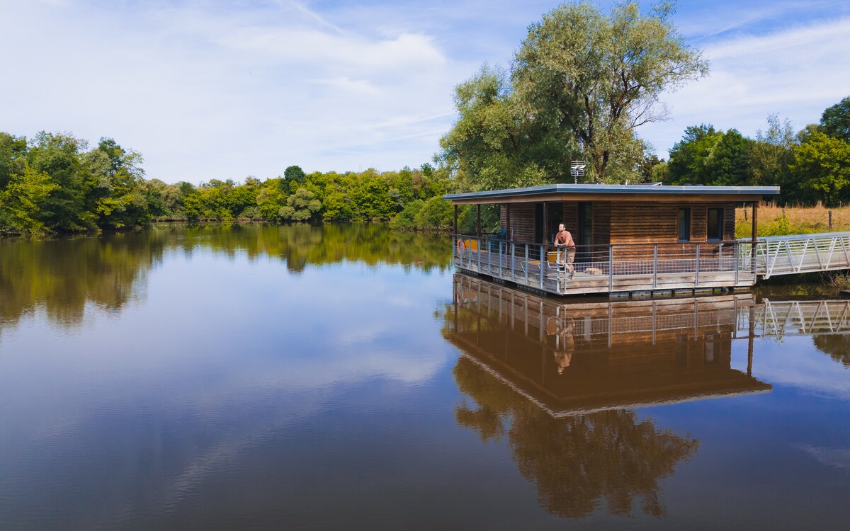 A modern lodge is situated on a calm water body, surrounded by lush green trees. The wooden structure features a spacious deck extending over the water, providing serene views. The tranquil environment reflects the lodge and nature, enhancing the peaceful setting.