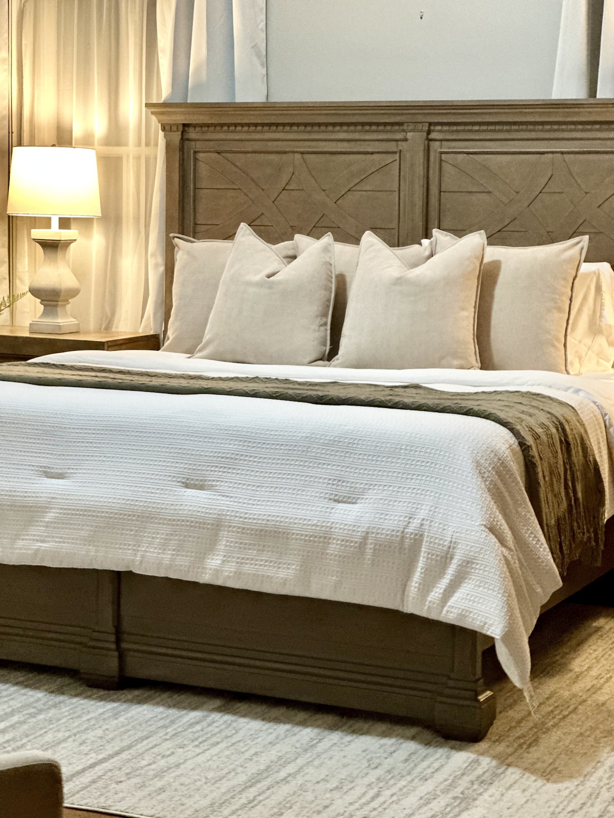 A spacious bed is dressed in soft white linens, complemented by decorative pillows in neutral tones. A bedside lamp provides gentle illumination, creating a calming environment. The wooden headboard features a subtle, carved design, contributing to the room's overall charm.