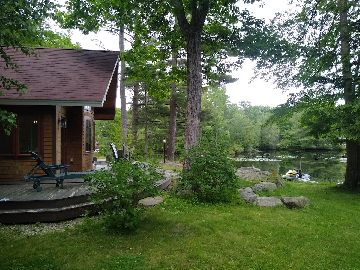 A lakeside view shows a cabin surrounded by greenery, with a wooden deck leading to the shoreline. Large trees provide shade, and a tranquil body of water can be seen in the background. Several rocks are present along the water's edge.