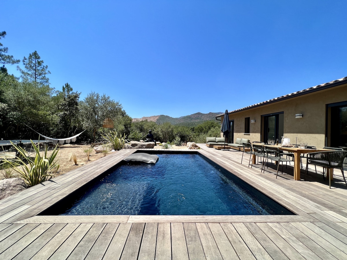 A serene pool area is framed by a wooden deck, featuring a dark blue swimming pool reflecting the clear sky. Surrounding vegetation includes trees and desert plants. A dining table and chairs are arranged nearby, with a sun umbrella providing shade.
