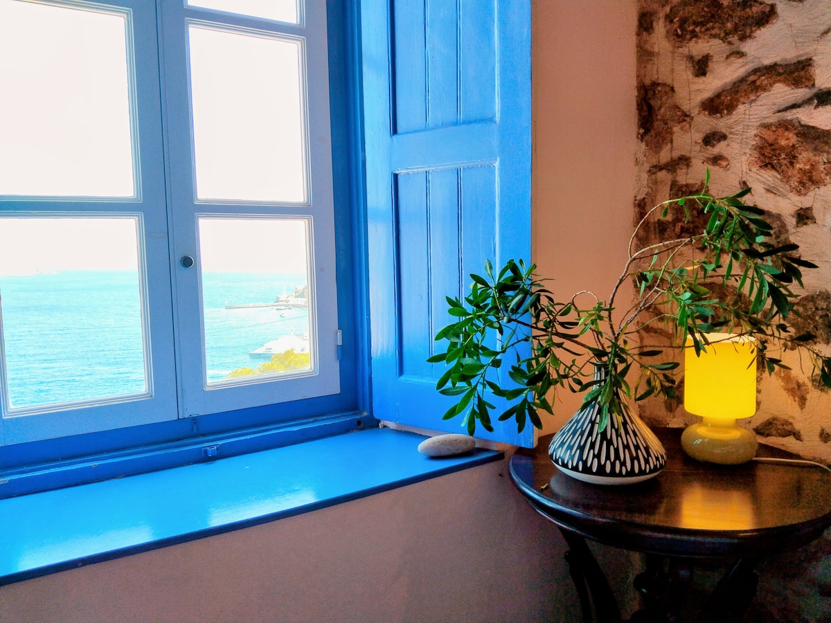 A blue window frames a scenic view of the sea, allowing natural light to fill the room. A decorative vase holds leafy branches on a small wooden table next to a soft-yellow lamp, complementing the stone wall background.
