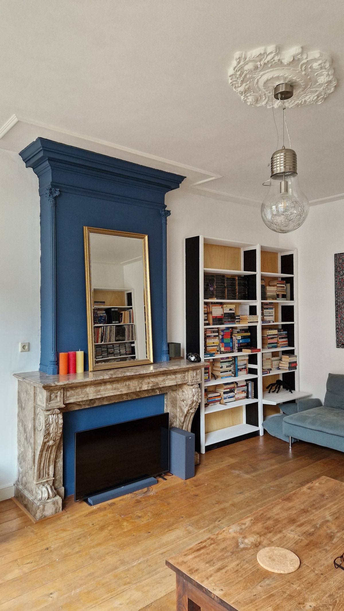 The image showcases a living area featuring a prominent blue accent wall behind a classic fireplace, adorned with a large mirror. A wooden coffee table is positioned in front of a comfortable seating area, complemented by shelves filled with books, enhancing the cozy atmosphere.