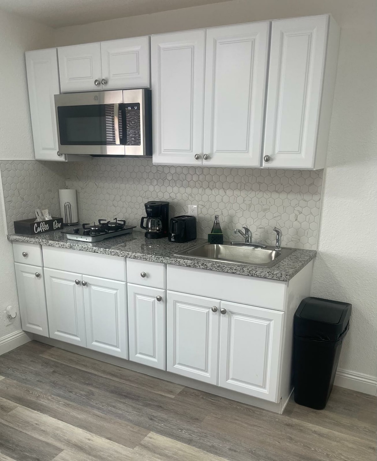 A modern kitchen area is presented with white cabinetry and a speckled countertop. A microwave is mounted above a gas stove. A coffee maker and a sink are included, along with a small black trash can. A tiled backsplash adds a subtle texture.
