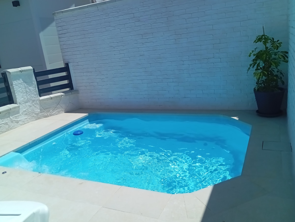 A private pool features clear blue water, surrounded by light-colored tile. The pool's shape is defined, with potted greenery located nearby. The white brick walls create a calm and relaxing atmosphere in this outdoor space.