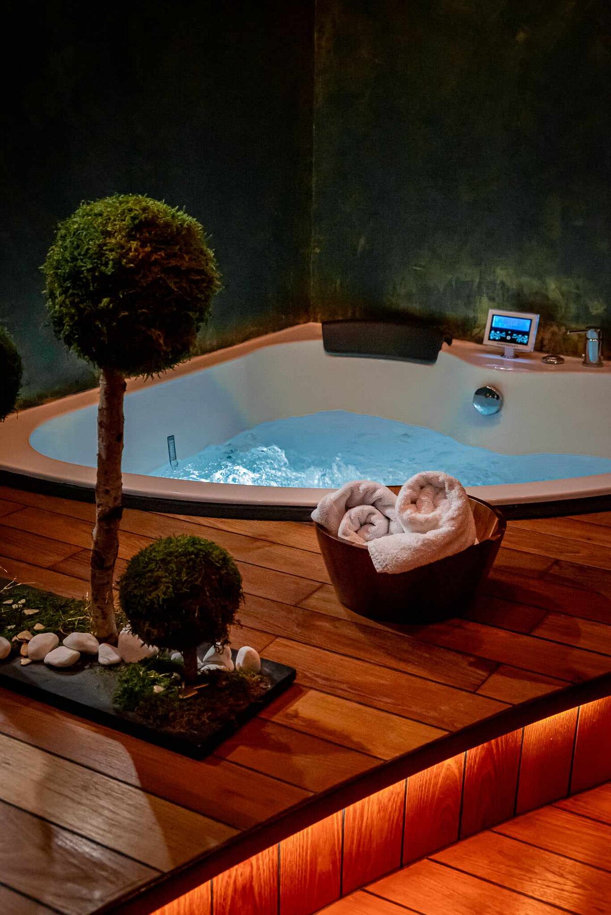 A private spa area features a soothing whirlpool bath with illuminated water, surrounded by wooden decking. Soft, rolled towels are placed in a wooden basket near a decorative arrangement of small stone and greenery.