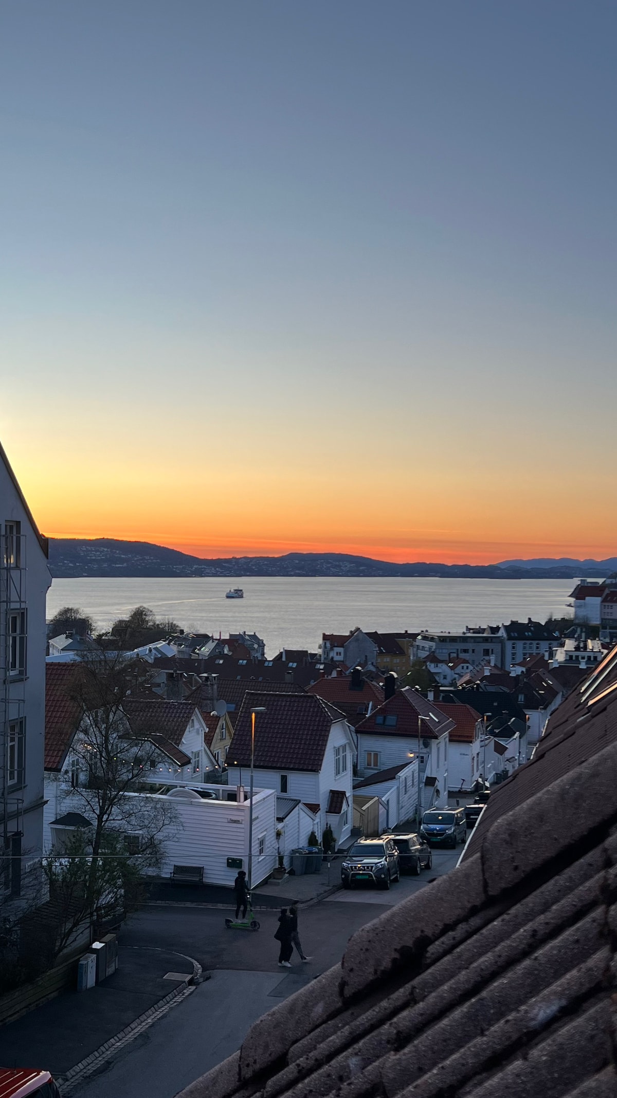 A panoramic view of a coastal town at sunset is displayed, with rooftops and streets visible in the foreground. The colors of the sky transition from orange to deep blue, reflecting on the water. Hills are seen in the background, adding depth to the picturesque landscape.