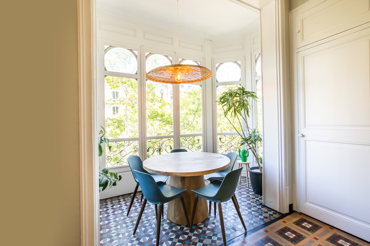 A dining area features a round wooden table surrounded by four modern chairs. Large windows allow natural light to fill the space, framed by decorative wrought iron. Green plants are placed in the corners, enhancing the fresh ambiance of the room.
